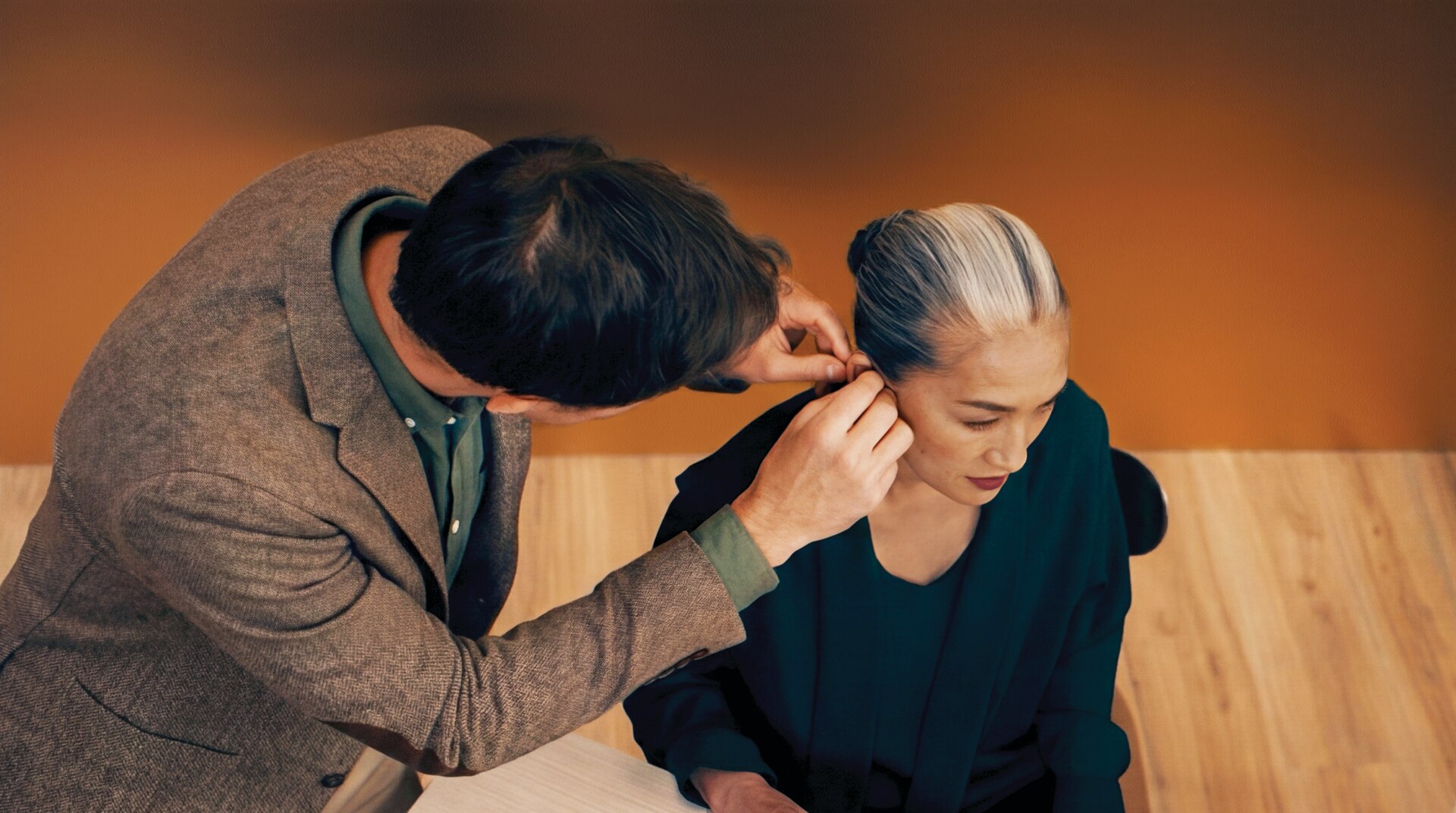 Man in a brown blazer leans over a seated woman with gray hair, helping adjust a device near her ear in a warm-toned room with a wooden floor.