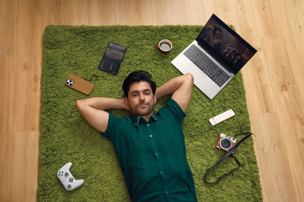 Man in a green shirt lying on a green rug on a wooden floor, with a laptop and camera nearby among other gadgets.