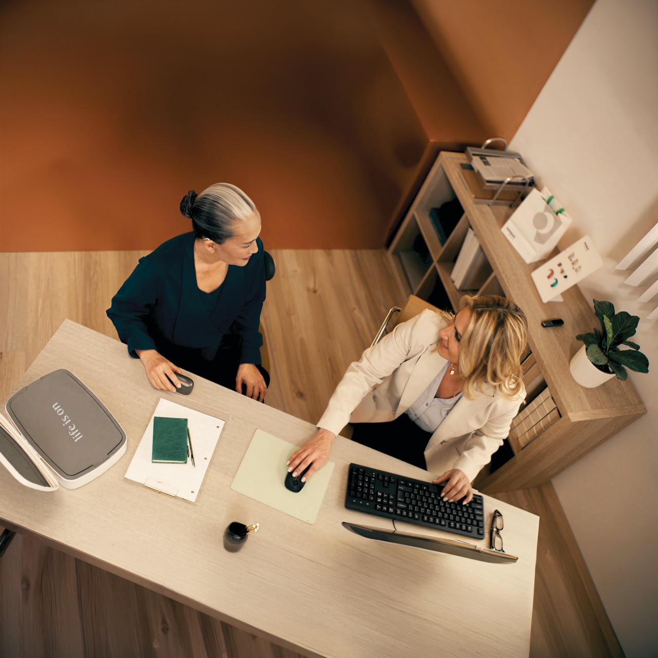 Two women are sitting at a wooden table in a brightly lit office. 