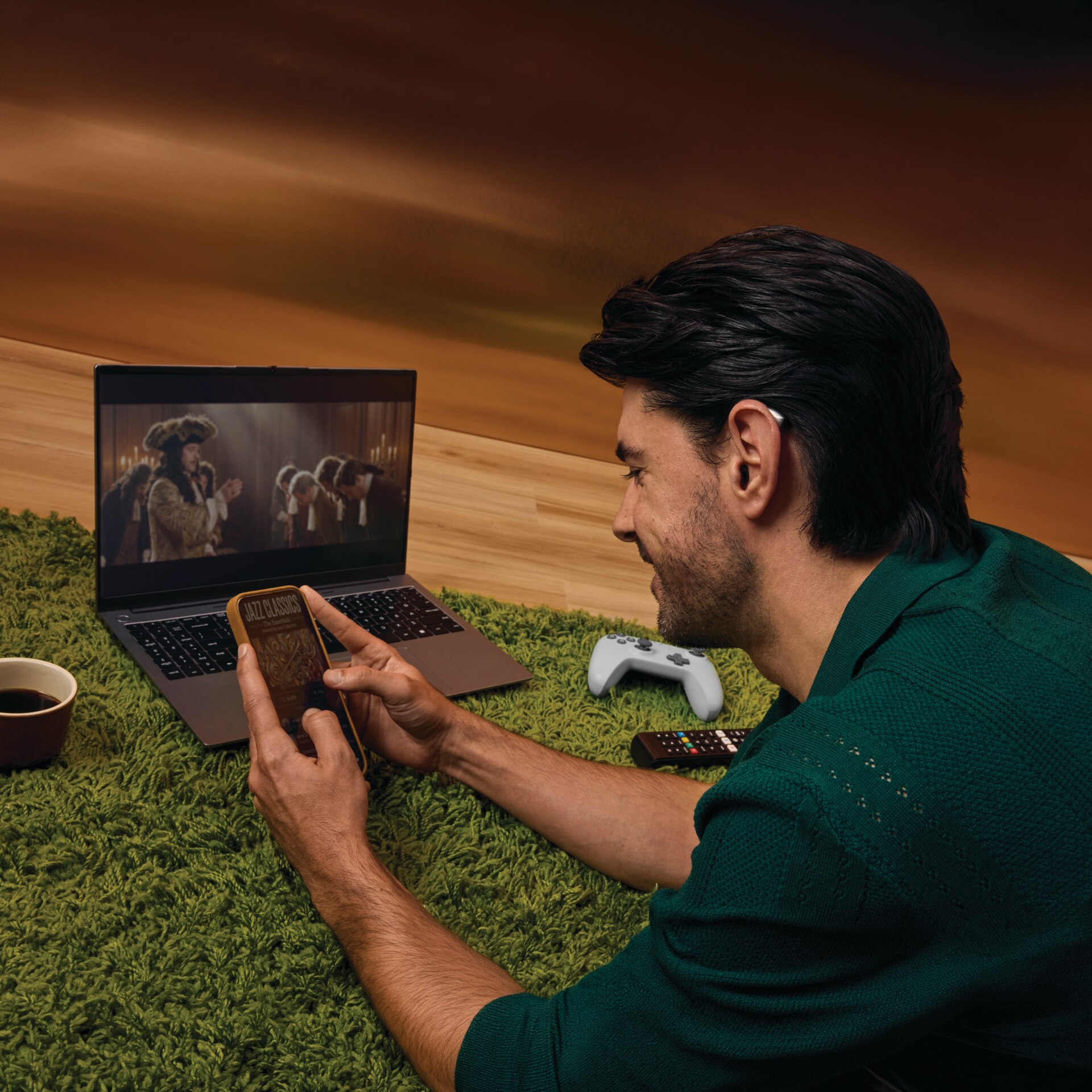 Person using a smartphone with a laptop and gaming controller nearby, illustrating hearing aid connectivity across everyday devices