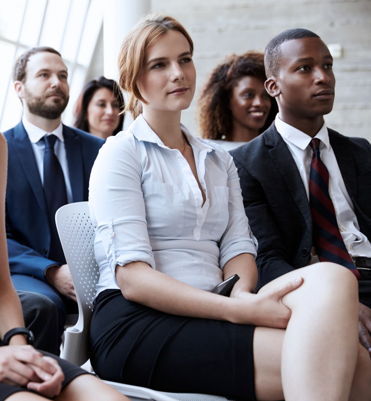 A man seated rows behind the speaker sits in a crowded auditorium watching a presentation.