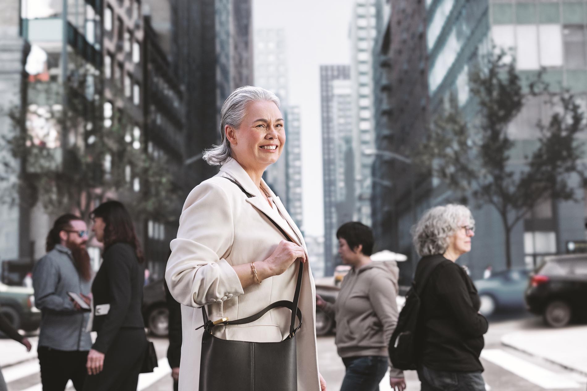 A smiling elderly woman crossing the street, wearing a light jacket and carrying a handbag