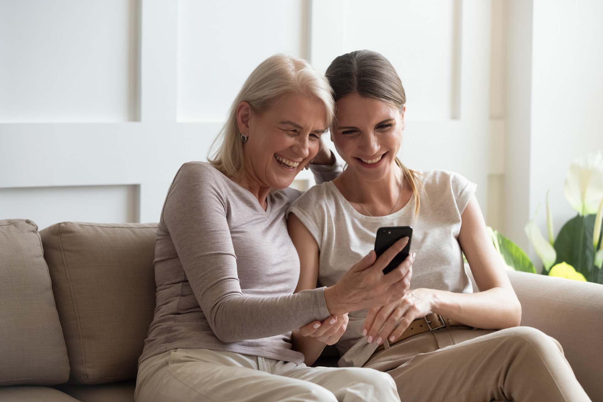 Mamma di mezza età e figlia adulta sedute sul divano di casa ridono guardando un video divertente sullo smartphone, madre anziana felicissima e figlia adulta si divertono usando un moderno cellulare