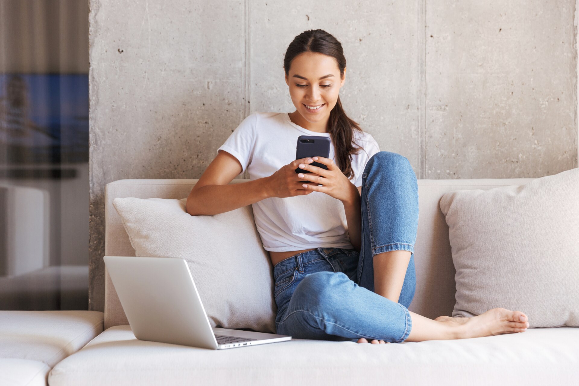 Giovane donna asiatica felice che utilizza il telefono cellulare mentre è seduta sul divano di casa con il computer portatile