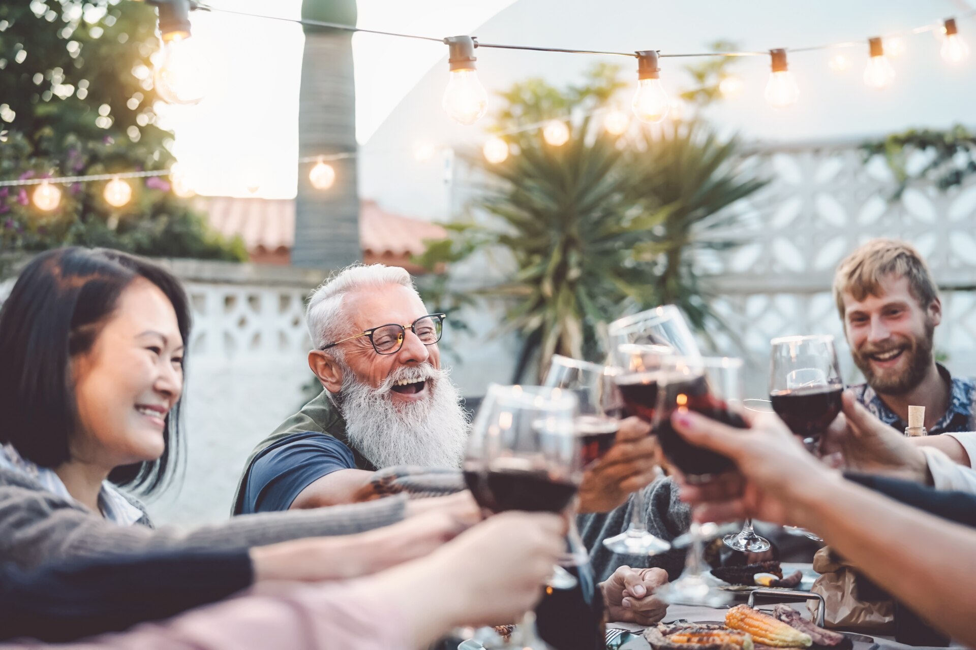 Famiglia felice che pranza e brinda con bicchieri di vino rosso all'aperto - Persone di diverse età ed etnie che si divertono in una cena con barbecue - Concetto di attività di fine settimana di giovani e anziani con genitori