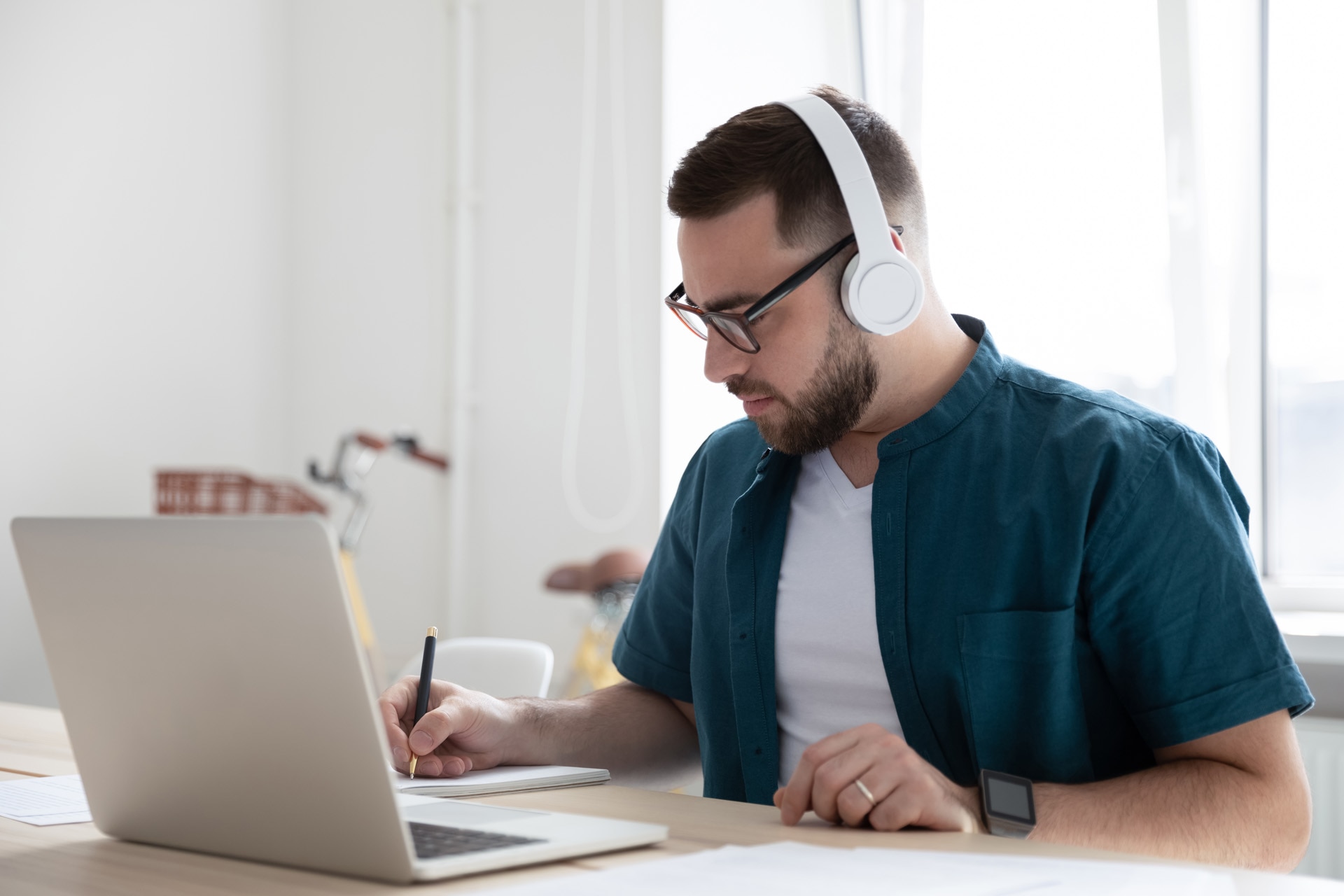 Focused young man businessman company worker employee in glasses wearing wireless headphones, watching educational webinar lecture seminar on laptop online, writing down notes in modern office.; Shutterstock ID 1653287650; purchase_order: -; job: -; client: -; other: -