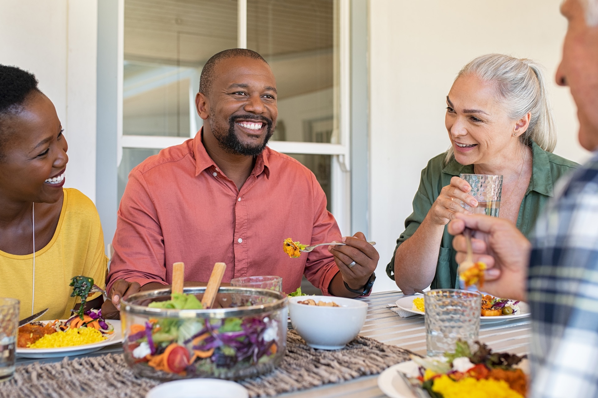 Gelukkige, lachende vrienden die samen thuis van een lunch genieten. Volwassen mensen met meerdere etnische achtergronden vieren een blijde gebeurtenis en eten gezond voedsel. Ouder echtpaar en Afrikaans echtpaar voeren gesprek tijdens maaltijd.