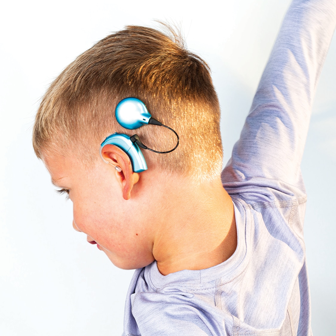 En ung gutt med kort hår, iført en lys lilla genser, løfter armen sin. Han har en høreapparat festet bak øret. Bakgrunnen er lys og enkel.