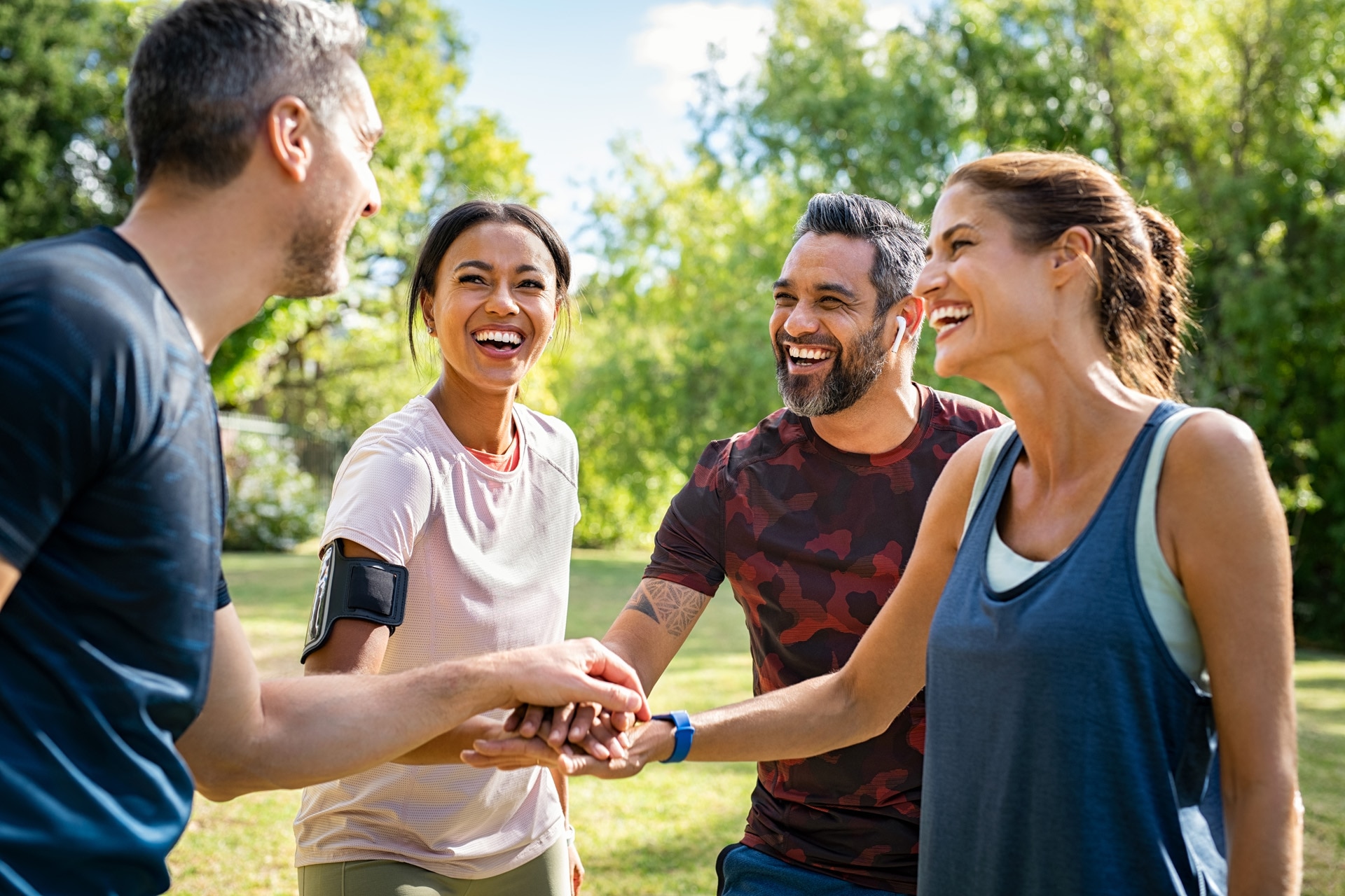 Laughing mature and multiethnic sports people at park. Happy group of men and women smiling and stacking hands outdoor after fitness training. Mature sweaty team cheering after intense training