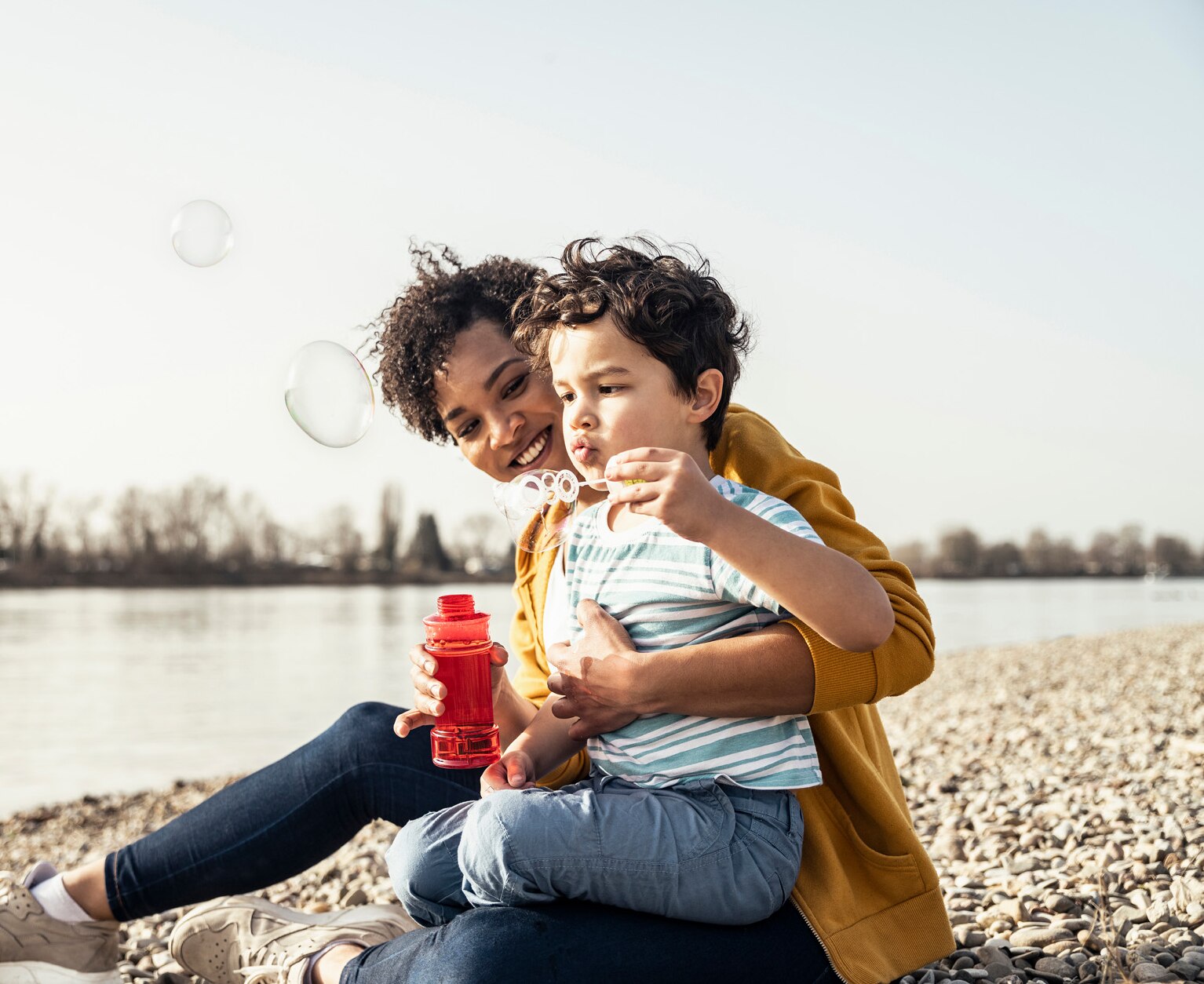 Boy playing with bubble wand while sitting on mother's lap during sunny day