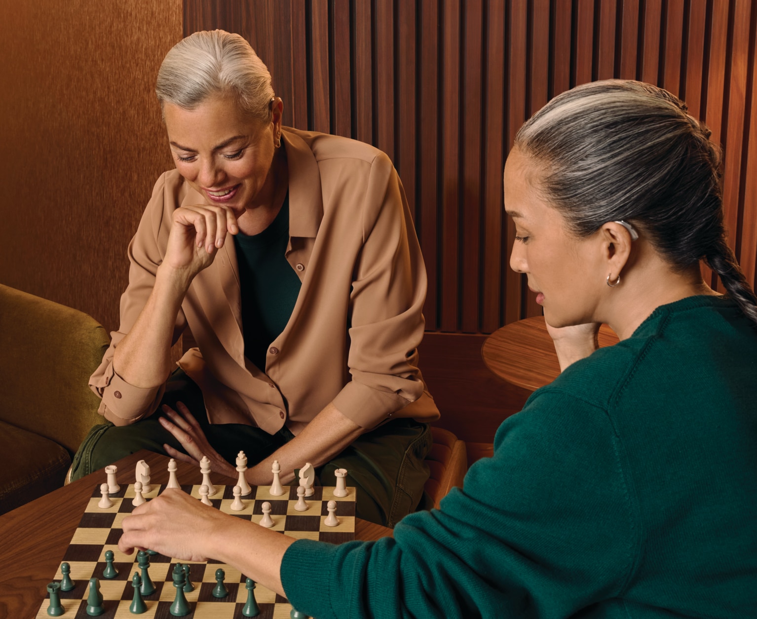 Two people play chess at a wooden table in a warm, wood-paneled room; one person with gray hair in a tan blazer leans over the board, while the other wears a green sweater and concentrates on the game, with a chessboard between them.
