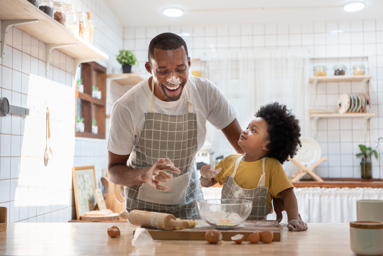 Cheerful smiling Black son enjoying playing with his father while doing bakery at home. Playful African family having fun cooking baking cake or cookies in kitchen together. Single Dad Lifestyle; Shutterstock ID 1950537220; purchase_order: -; job: -; client: -; other: -