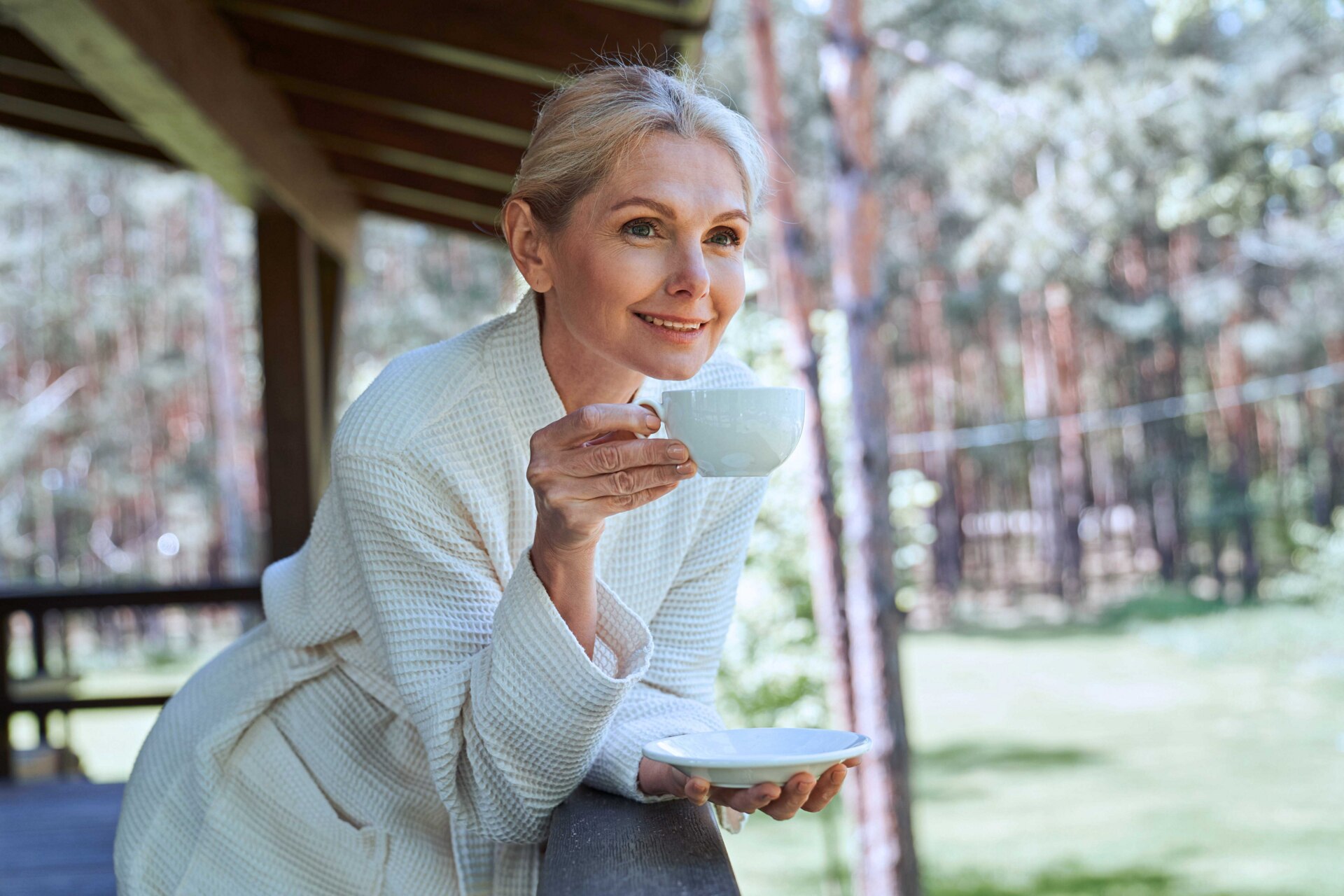 A woman leans against an outdoor patio railing while sipping a beverage and enjoying the view on a sunny day