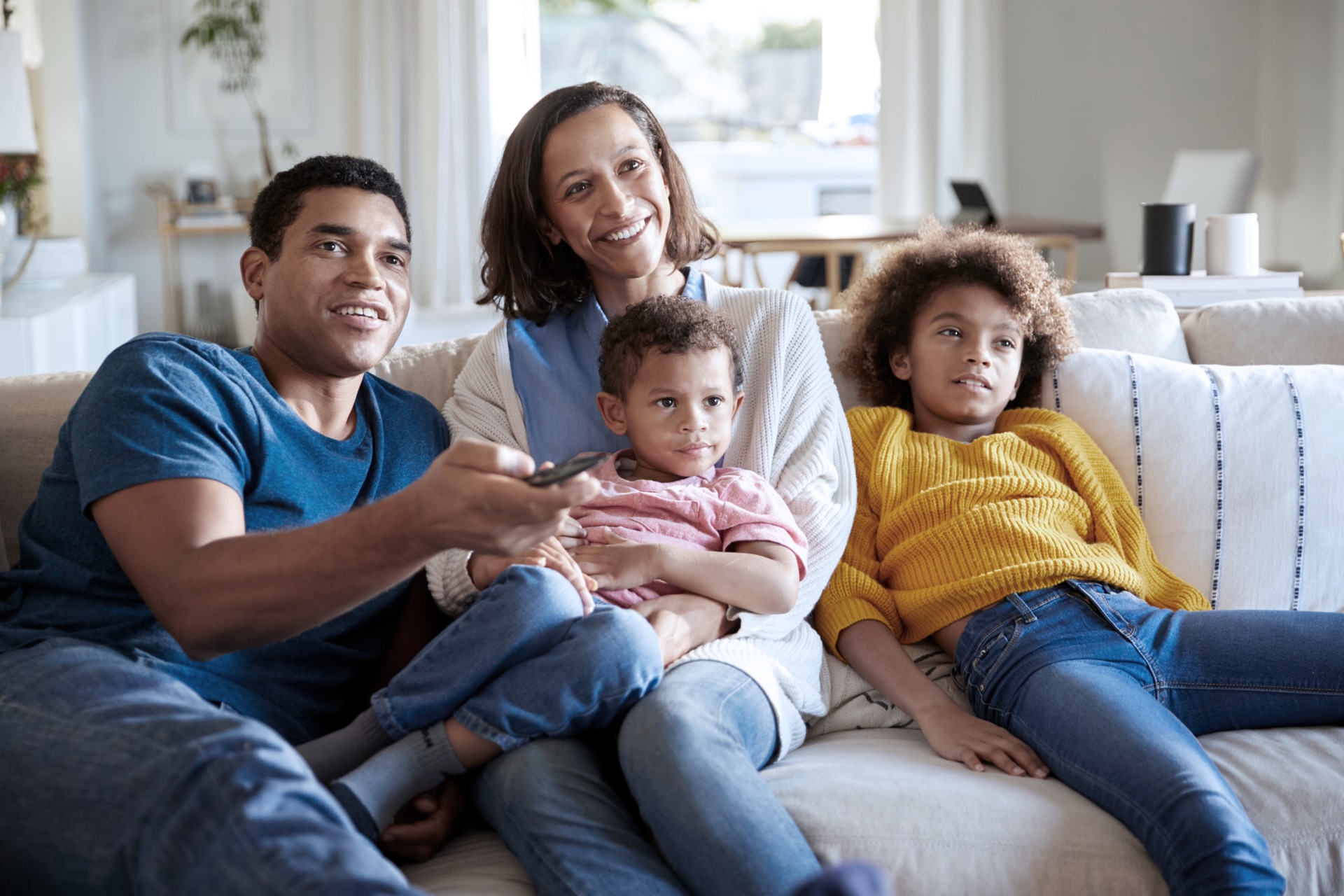 A happy family sits on the couch while the father points a remote control at the TV.