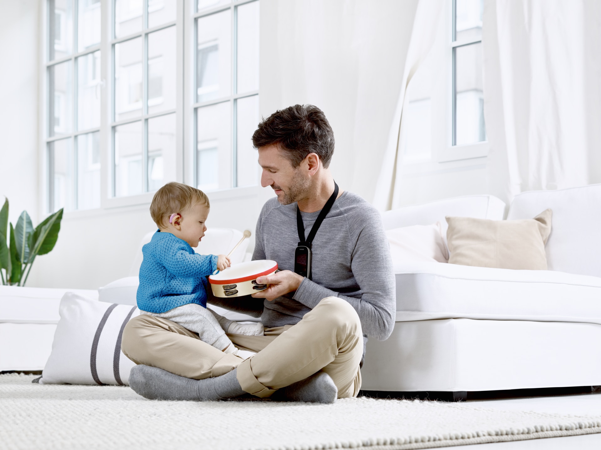 Toddler with a hearing aid sitting on his father's lap playing on a drum.