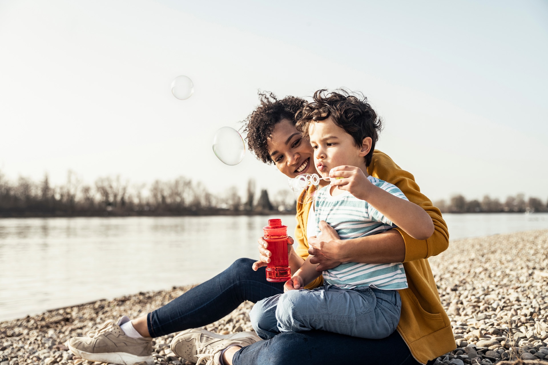 Boy playing with bubble wand while sitting on mother's lap during sunny day