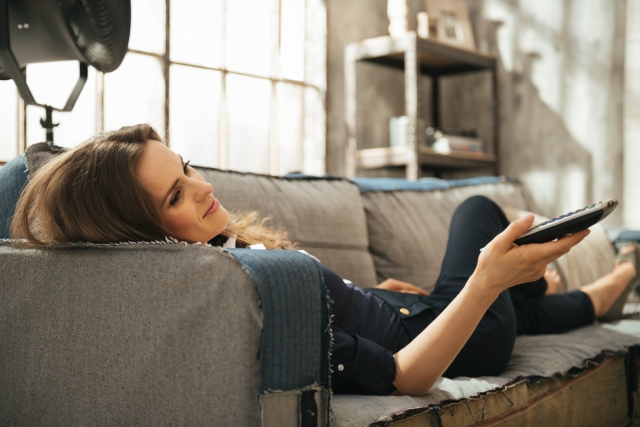 Avslappnad ung brunett som ligger i soffan och tittar på TV i en loftlägenhet. Stads-chic inredning och fönster.