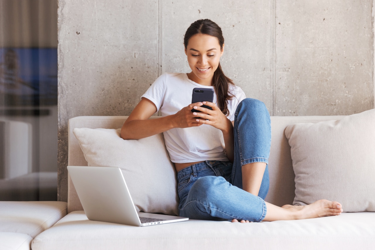 Glad, ung asiatisk kvinna som använder sin mobiltelefon medan hon sitter med en bärbar dator hemma i sin soffa