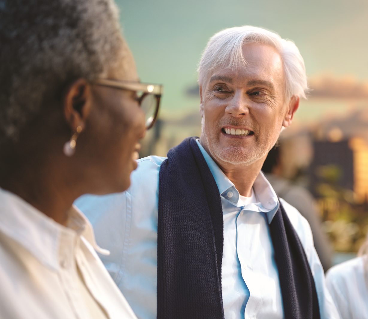 A group of casually dressed adults are conversing outdoors in an urban plaza, with tall buildings visible in the background and a warm sunset glow in the sky. En grupp människor som ler