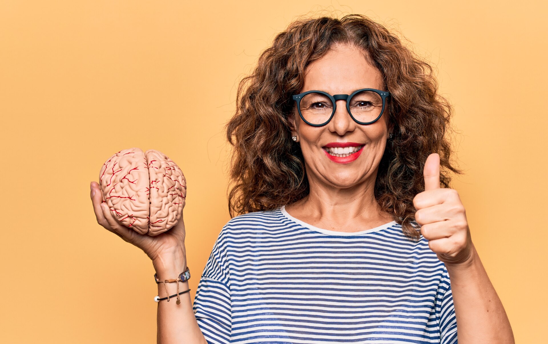 1808138263 Middle age brunette woman holding brain as mental care and memory health over pink background smiling happy and positive, thumb up doing excellent and approval sign