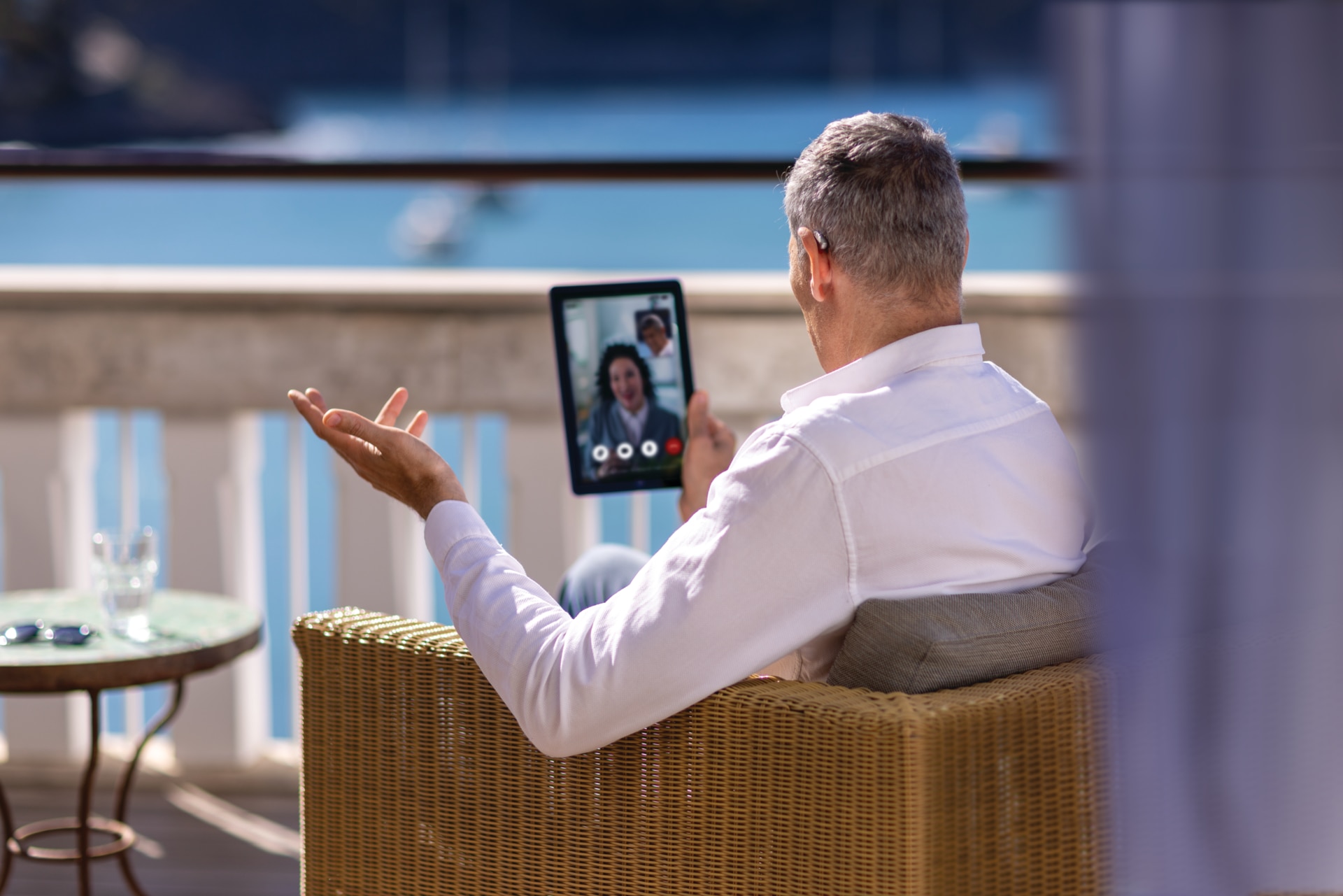 An elegant man sitting on a hotel balcony with a tablet connecting to a hearing care specialist via remote support