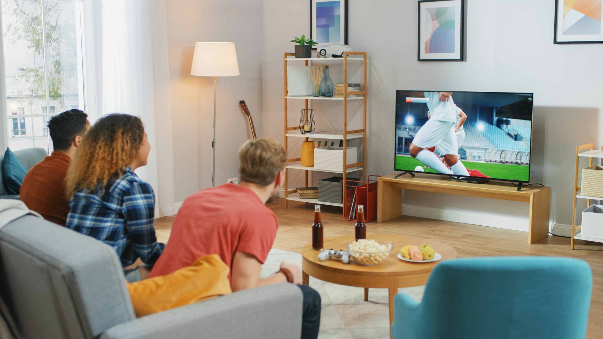 Three Sports Fans Sitting on a Couch in the Living Room Watch Important Soccer Match, Worry and Cheering For their Team. Bright Cozy Apartment with Friends Eating Snacks and Having Fun.