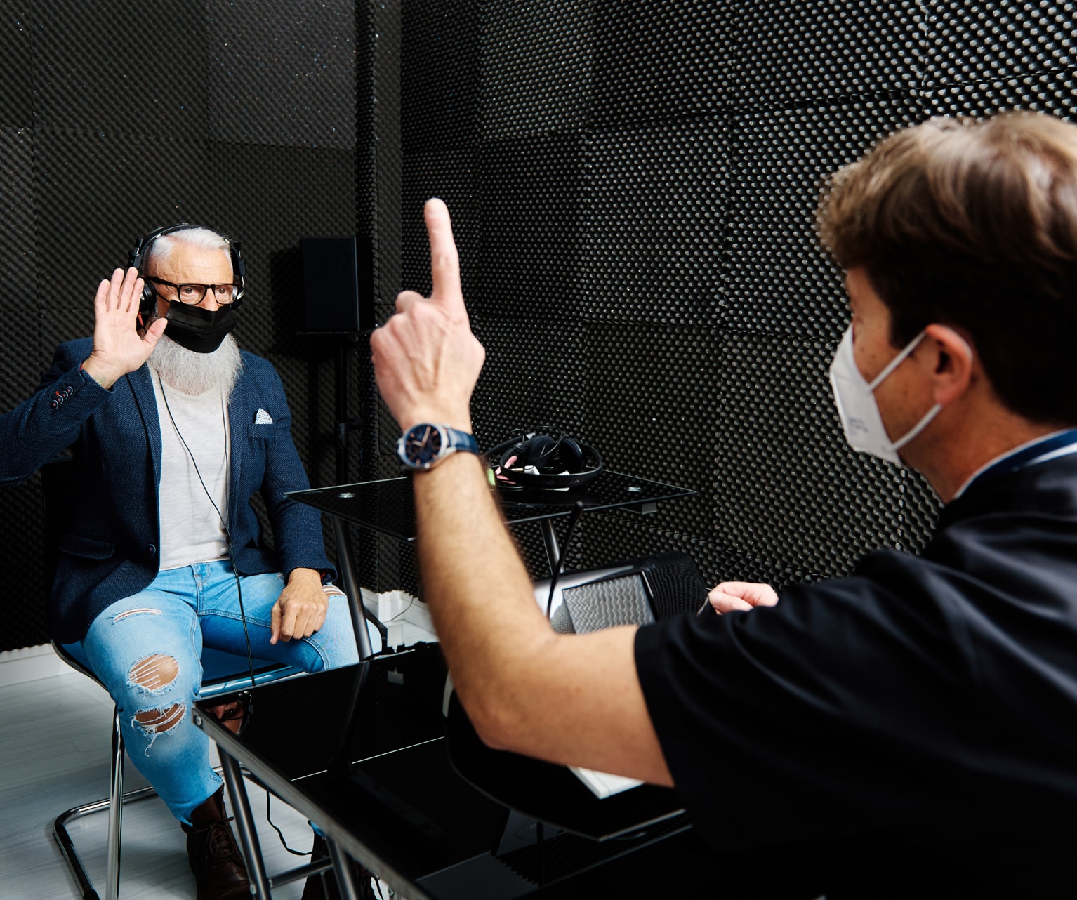Aged male in headphones and mask sitting in room with doctor conducting audiological hearing test.