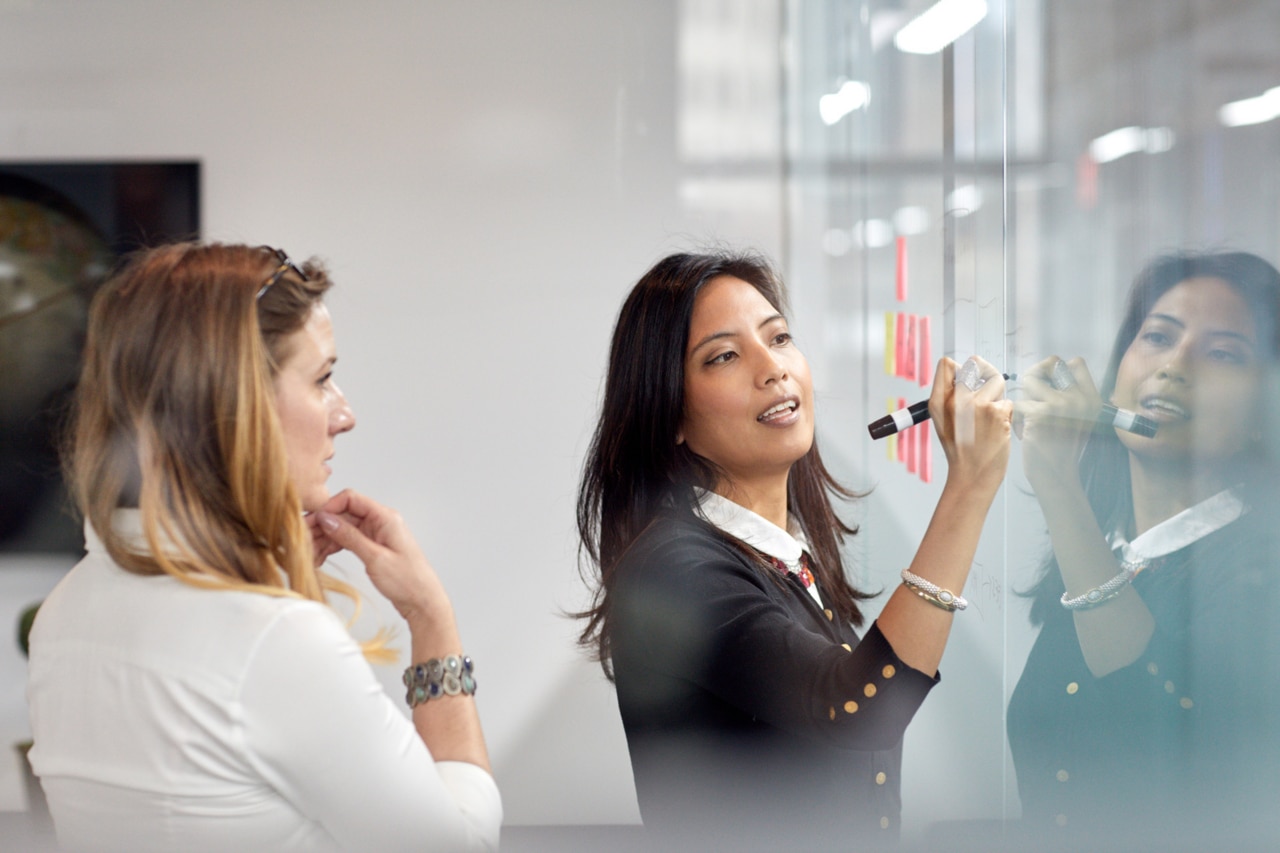 Businesswoman looking at female colleague writing on adhesive notes in office