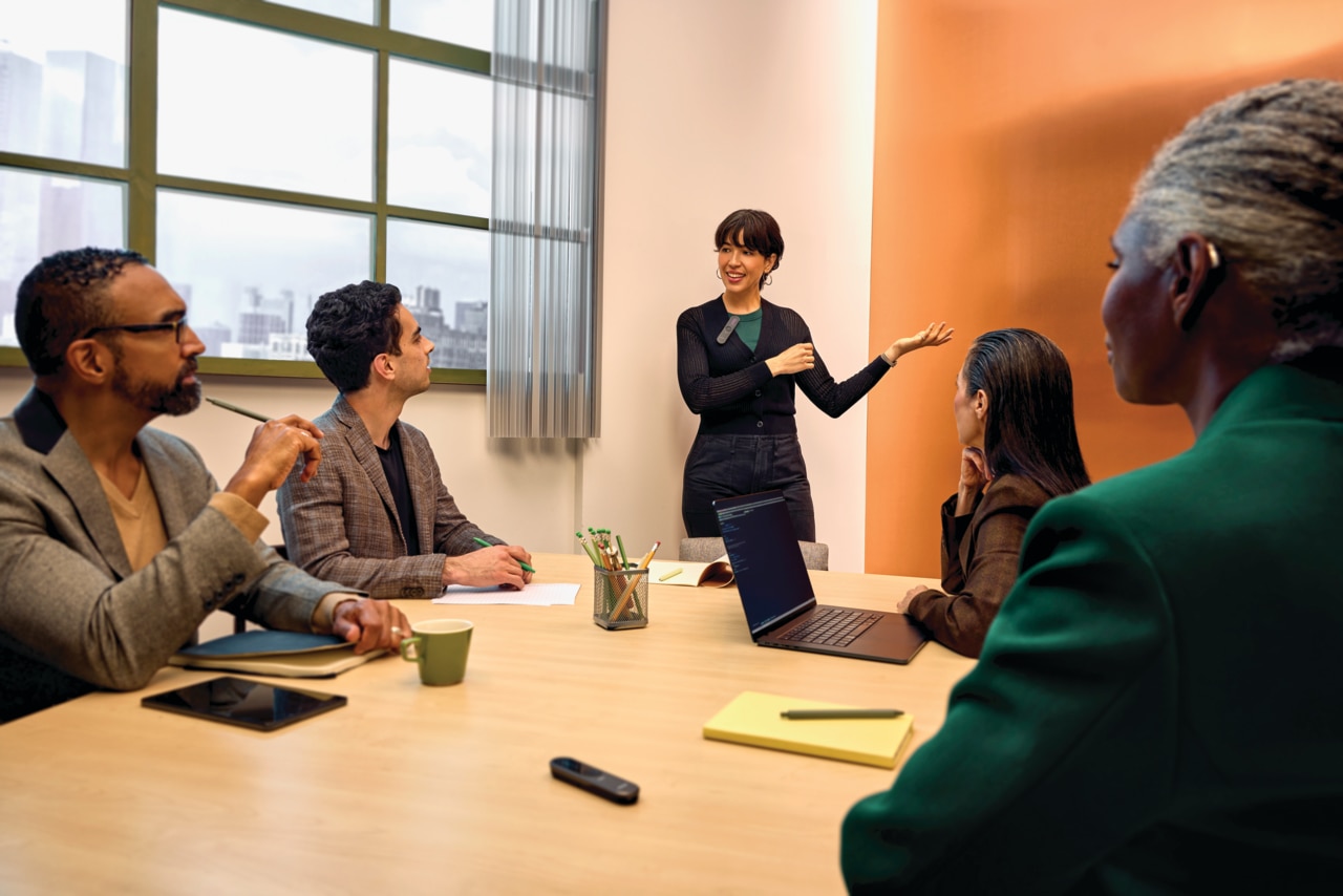 A group of people participating in a meeting, sitting at a round table, listening to a female speaker