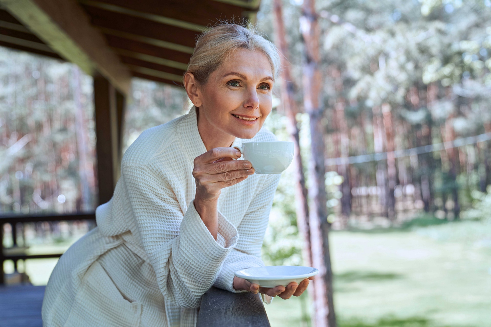 A woman leans against an outdoor patio railing while sipping a beverage and enjoying the view on a sunny day