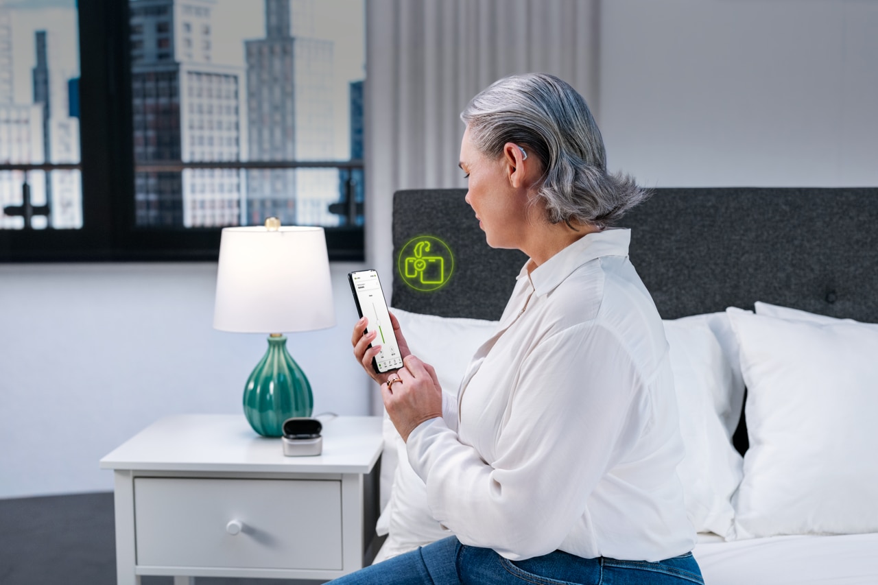 An elderly woman sitting on a bed in a brightly lit bedroom looking at her smartphone