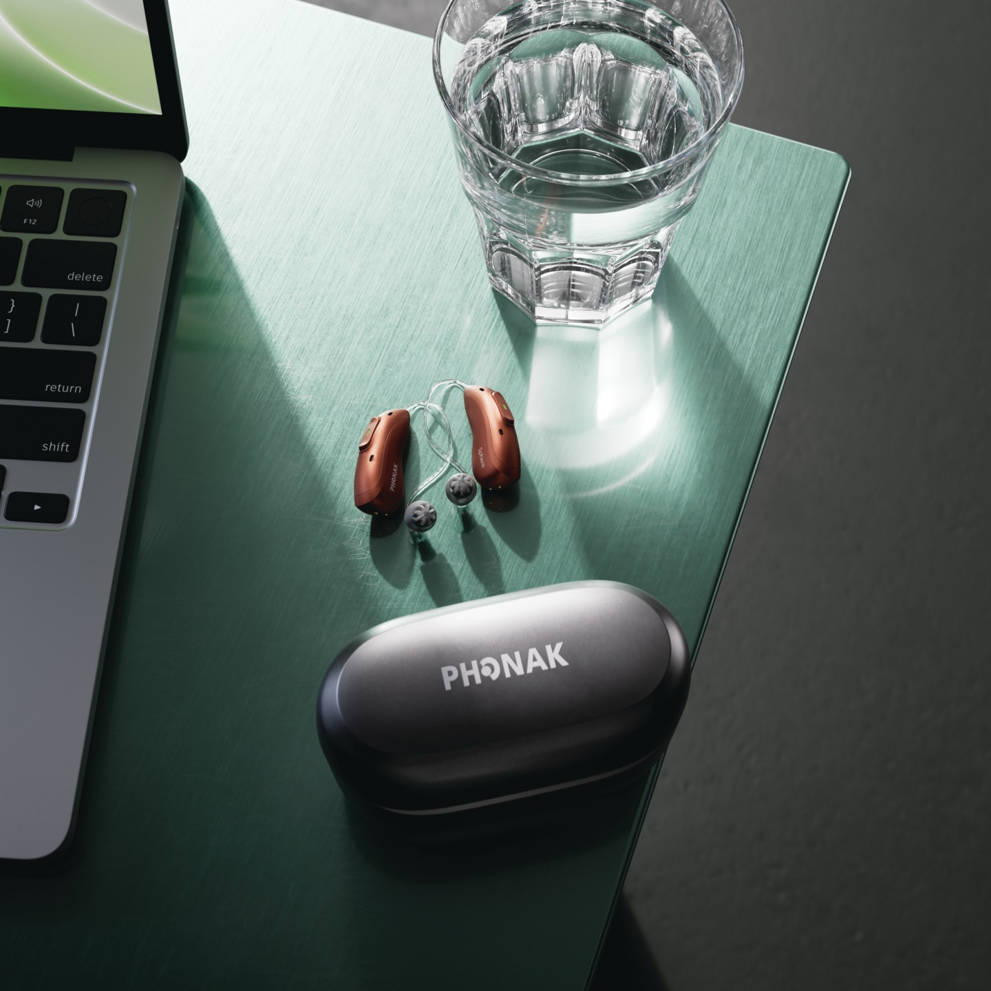 Green desk with a laptop edge on the left, a glass of water in the center, and a black PHONAK case with two brown earbuds nearby.