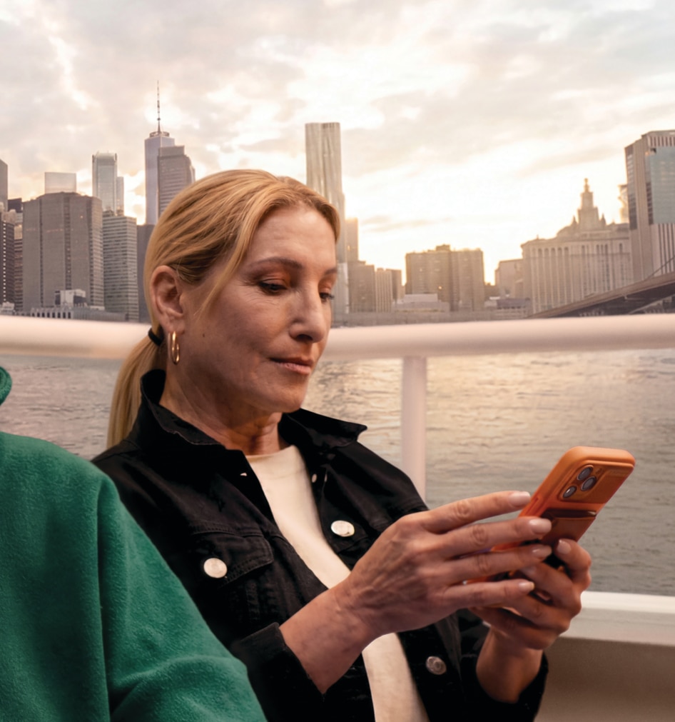 Two women sit on a boat by the water with a city skyline at sunset in the background. The woman in the foreground wears a green fleece and smiles softly at the camera, while the woman behind her in a black jacket looks down at a smartphone.