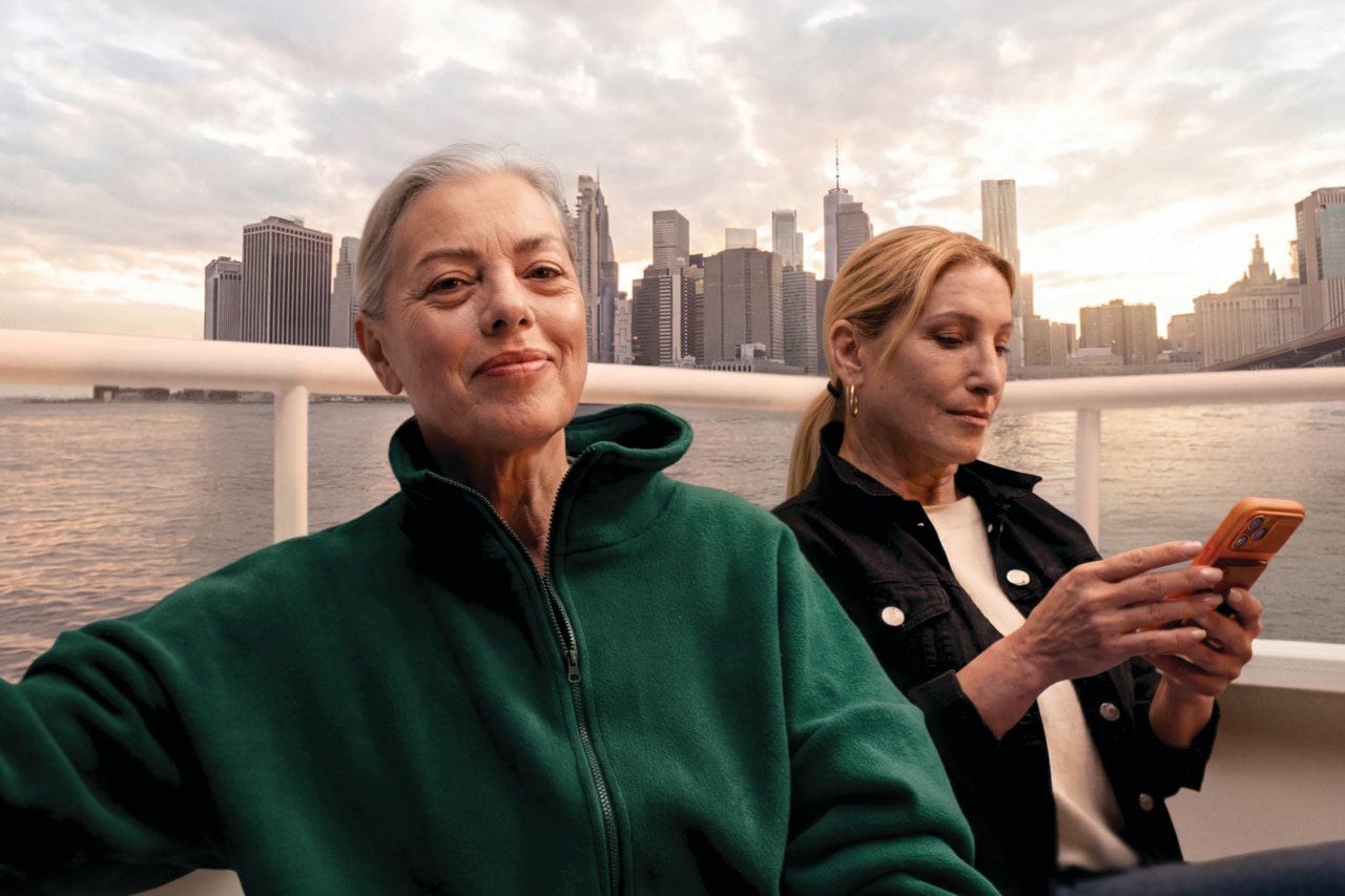 Two women sit on a boat by the water with a city skyline at sunset in the background. The woman in the foreground wears a green fleece and smiles softly at the camera, while the woman behind her in a black jacket looks down at a smartphone.