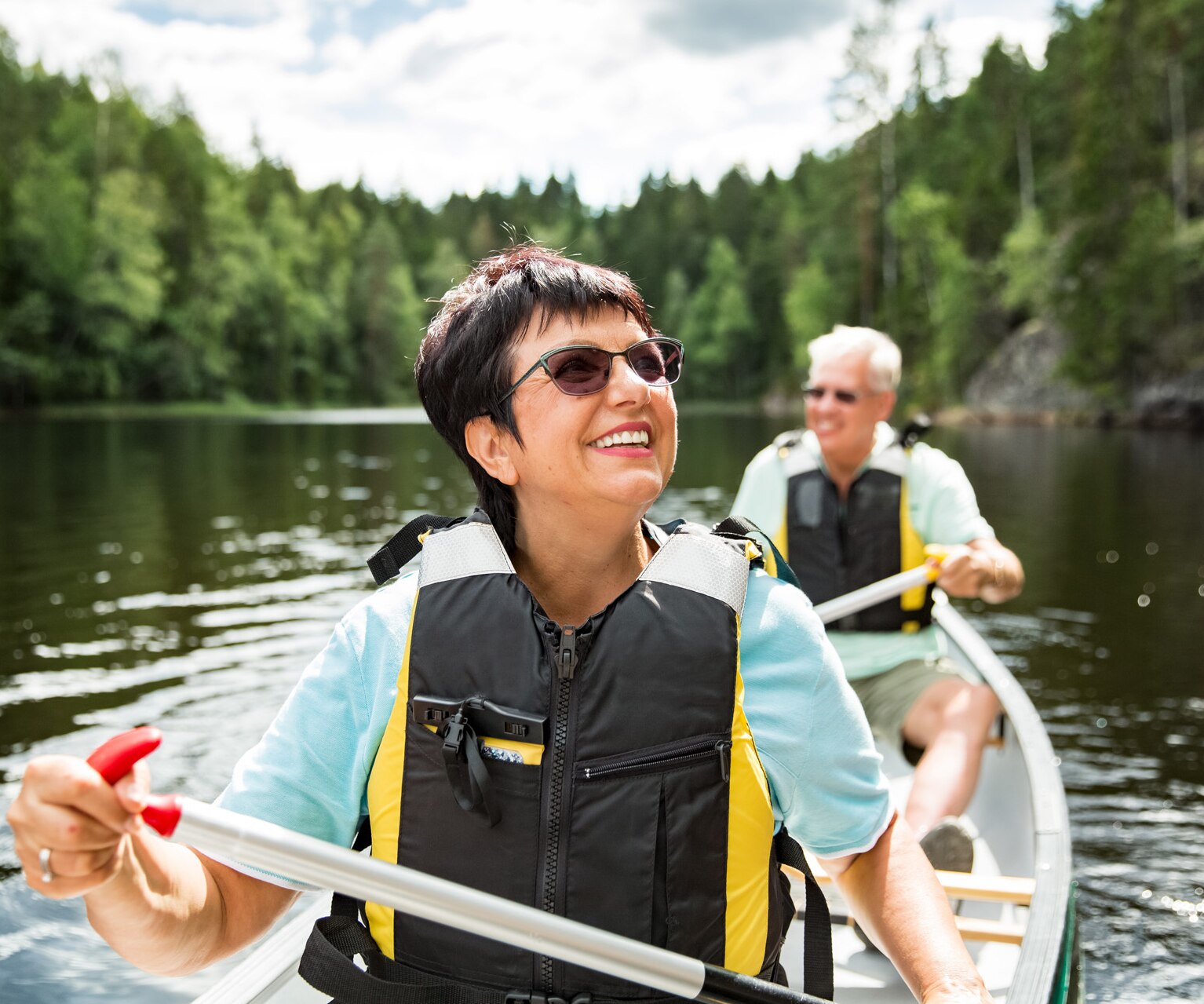 Happy mature couple in life vests canoeing in forest lake. Sunny summer day. Tourists traveling in Finland, having adventure.