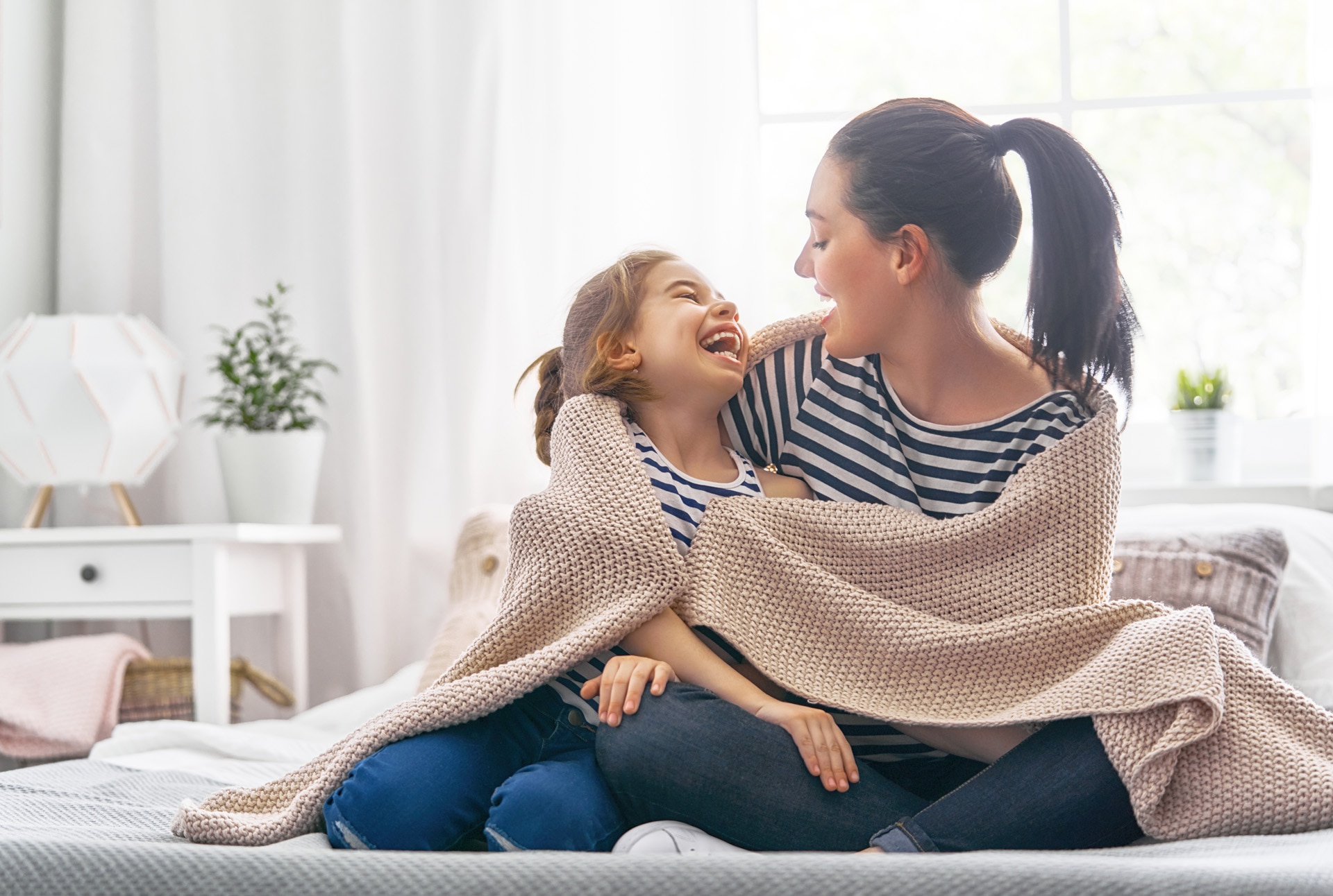 A nice girl and her mother enjoy sunny morning. Good time at home. Child wakes up from sleep. Family playing under blanket on the bed in the bedroom.; Shutterstock ID 1066576061; purchase_order: -; job: -; client: -; other: -