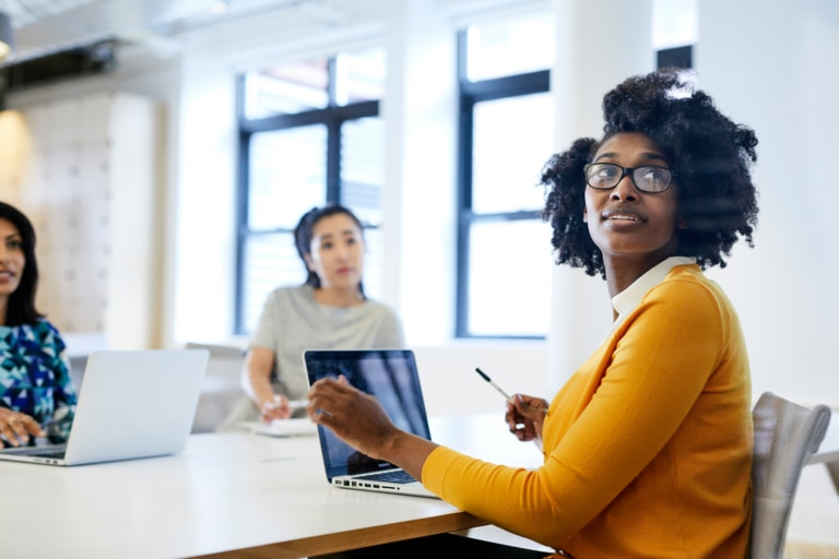 female wearing mustard colored shirt working on her laptop at a desk with two peers