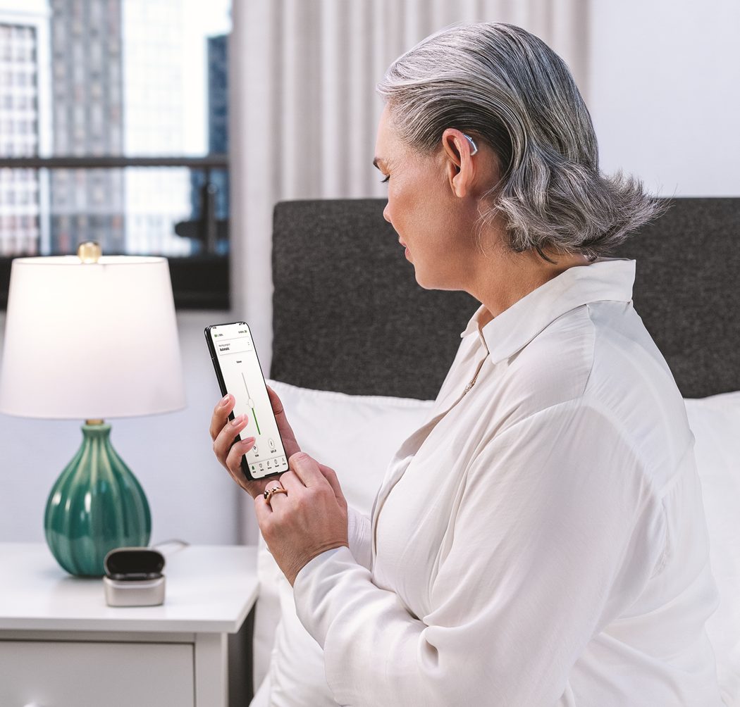 An elderly woman sitting on a bed in a brightly lit bedroom looking at her smartphone