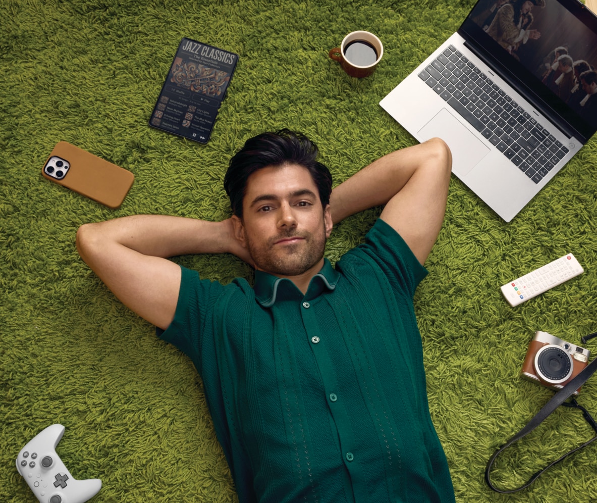 Man in a green shirt lying on a green rug on a wooden floor, with a laptop and camera nearby among other gadgets.