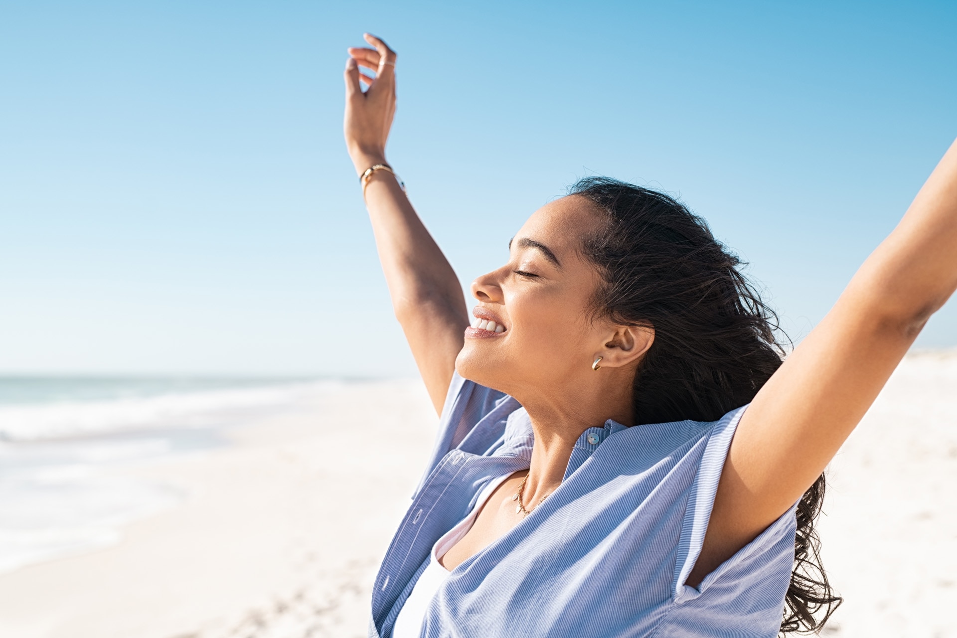 Smiling latin hispanic woman stretching hand and relaxing on beach. Woman breathing deeply at seaside with eyes closed. Happy woman standing on the beach and enjoy the sun tan with arms outstretched.; Shutterstock ID 2134545737; purchase_order: -; job: -; client: -; other: -